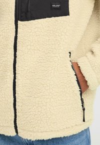 Beige sherpa fliisjakki mustade tõmblukudetailidega, rinna tasku ja käsi osaliselt küljetaskus.