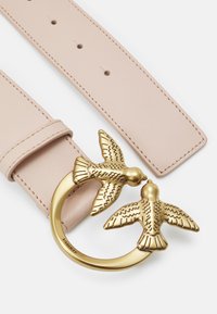 Gaiši rozā ādas josta ar apaļu zelta sprādzi, kurā attēlots divas smalki izstrādātas putni. Jostas lentei ir redzamas šuves un regulēšanas caurumi.