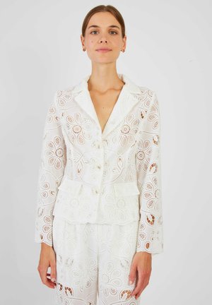 Kobieta w białym koronkowym blezerze i dopasowanych spodniach z kwiatowymi wycięciami, stojąca na tle jednolitego jasnego tła.