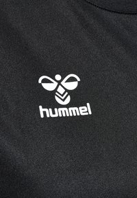 Musta urheilupaita, jossa on suuri valkoinen logo, jonka keskellä on mehiläisen muotoilu ja sen alla teksti "hummel". Pehmeä kankaan tekstuuri.