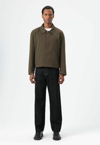 Olijfgroene cropped jas met kraag, frontale knoopsluiting, gecombineerd met zwarte rechte jeans en zwarte stevige schoenen.