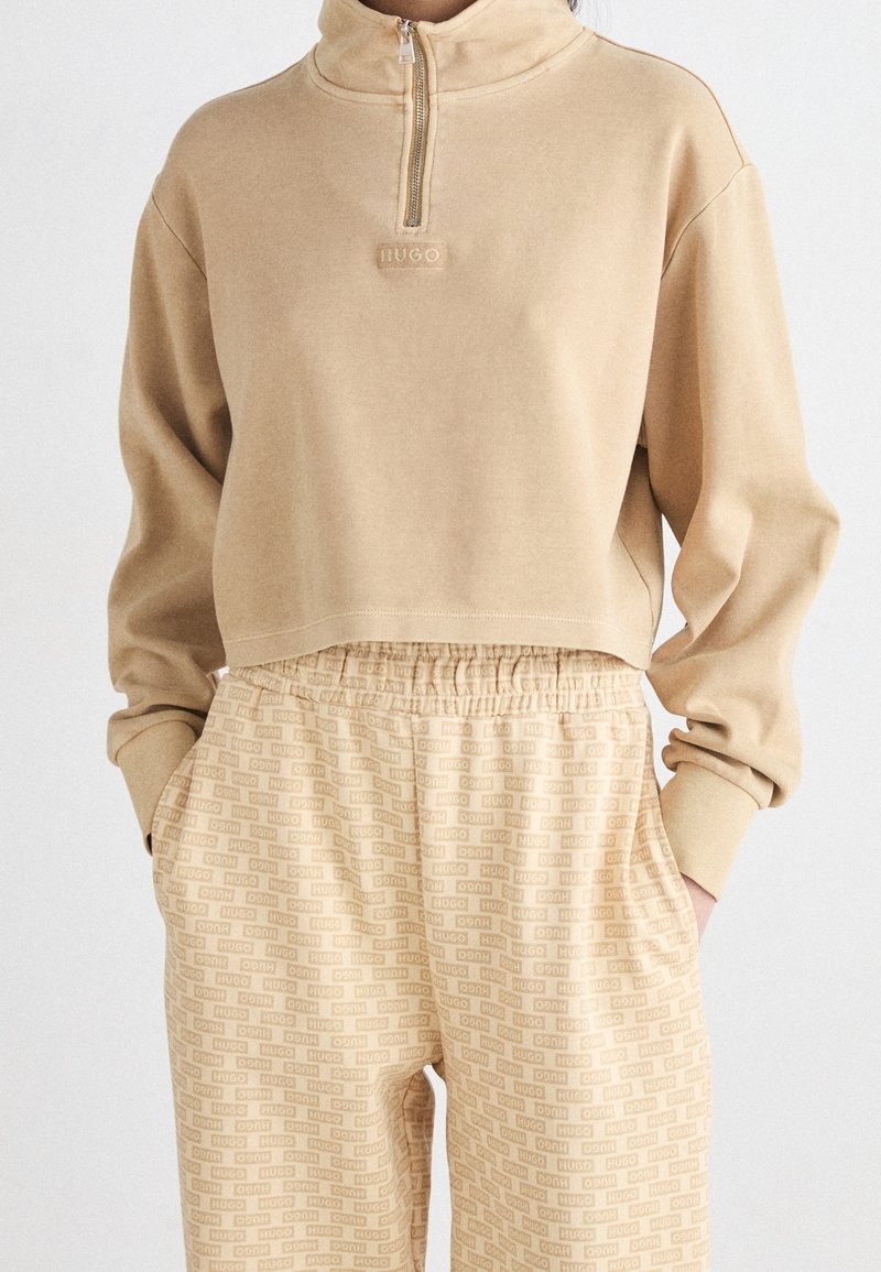 Sweat-shirt court beige avec une fermeture éclair au quart et pantalon beige à taille élastique assorti avec un motif de texte intégral.