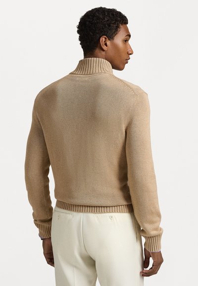Beige stickad tröja med hög krage, ribbade ärmslut och nederkant; har ett strukturerat mönster och en avslappnad passform, ihop med ljusa byxor.