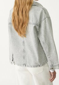 Šedá džínová bunda s volným střihem, se špičatým límcem, knoflíkovými manžetami a švy na zádech, nošena přes bílé kalhoty.