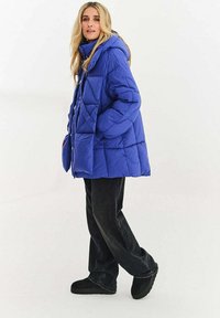 Přistřižená modrá prošívaná bunda s kapucí, velkým kosočtvercovým prošíváním, bočními kapsami a volným střihem, doplněná černými širokými kalhotami a černými botami.