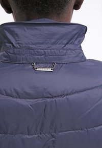 Vadderad marinblå jacka med hög krage, texturerad tyg och metallic logotypdetalj på nackområdet, säkrad med kedjedetalj.