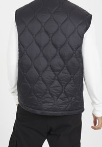 Black quilted vest with a rounded pattern, featuring a high neckline and a lightweight fabric. Worn over a white long-sleeve shirt.