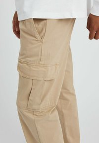 Pantalons cargo beiges à coupe décontractée, avec des poches latérales, des détails cousus et un tissu doux et texturé. Seule la partie inférieure est visible.