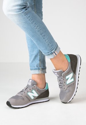 Person iført grå og turkise New Balance-sneakers med lyseblå, opsmøgede jeans, stående på en ensfarvet hvid overflade.