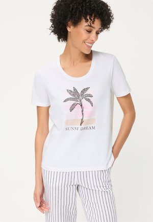 Lächelnde Frau mit lockigem Haar, die ein weißes T-Shirt mit einem Palmenmotiv und dem Schriftzug "Sunny Dream" trägt, kombiniert mit weißen, gestreiften Hosen.