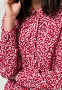 Femme portant une robe rouge avec des motifs floraux blancs et noirs, main levée près de son visage, et cheveux sombres jusqu'aux épaules visibles.