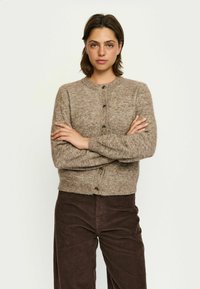 Rjav teksturiran kardigan s krožnim ovratnikom in gumbi spredaj, oblečen čez temno žametne hlače. Pletenina izgleda mehka in topla.