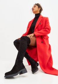Botas de ante negras hasta la rodilla con tacón cuadrado, combinadas con un abrigo largo rojo brillante sobre un top negro de cuello alto.
