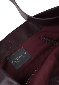 Interno di una borsa tote in pelle borgogna con foderatura in tessuto, un'etichetta con la scritta "TUCANO MILANO 1985" e un dettaglio con tasca con zip.
