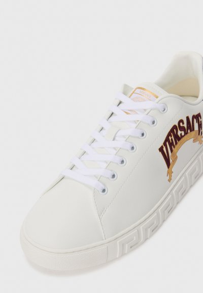 Adidași din piele albă cu cusături tonale, având un logo Versace ridicat, de culoare maro, pe lateral și o talpă din cauciuc texturat.