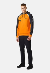 Bluza z kapturem o pomarańczowym przodzie i czarnych rękawach. Posiada kieszeń kangurkę, nadruki z logo oraz ściągacze przy mankietach. Połączona z czarnymi spodniami i butami.