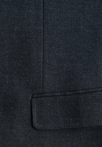 Tessuto di colore blu scuro con una trama texturizzata che presenta un sottile motivo intrecciato e una tasca a pattina rettangolare. Finitura liscia con un aspetto leggermente ruvido.