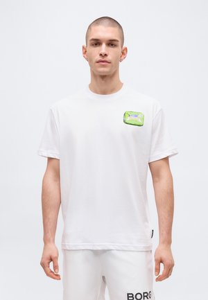 Νεαρός άνδρας με κοντά μαλλιά που φοράει λευκό μπλουζάκι με ένα μικρό πράσινο σημείο στο στήθος και λευκά σορτς με μαύρο κείμενο.