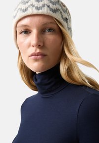 Młoda kobieta z blond włosami, nosząca białą i szaro-melanżową czapkę oraz granatowy sweter z wysokim kołnierzem na jednolitym tle.
