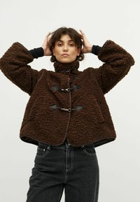 Donna che indossa una giacca in pile marrone a texture con chiusure a toggle, mentre posa con le mani dietro la testa e indossa jeans neri a vita alta.