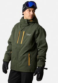 Grön softshelljacka med orange dragkedjor, flera fickor och huva. Svarta handskar och skidglasögon fulländar outfiten för vintersport.