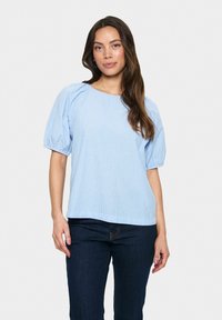 Blusa a righe blu chiaro con maniche a sbuffo, scollatura rotonda e vestibilità comoda, abbinata a jeans blu scuro. Tessuto di cotone liscio.