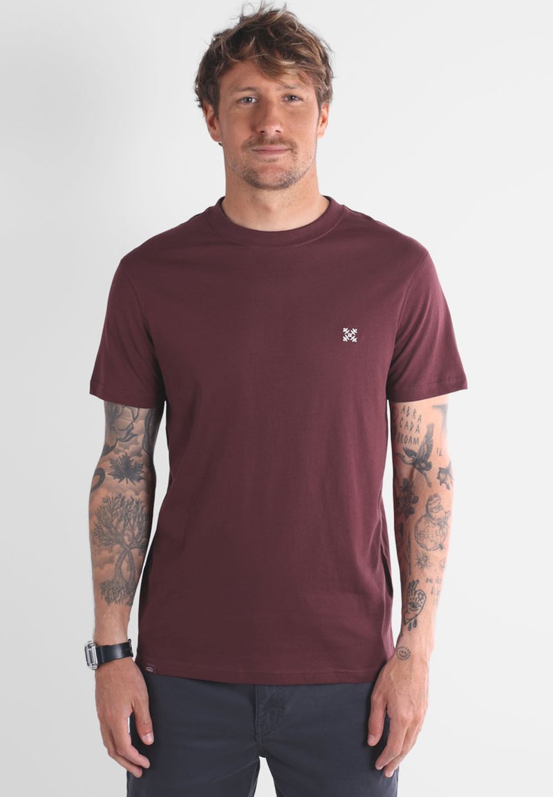 T-shirt en coton bordeaux avec un col rond, des manches courtes et un petit logo blanc brodé sur la poitrine gauche. Le modèle a des tatouages visibles.