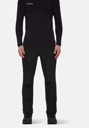 Persona che indossa un top a maniche lunghe nero Mammut, pantaloni neri con tasca con cerniera e scarponi da trekking neri, in piedi davanti a uno sfondo semplice.