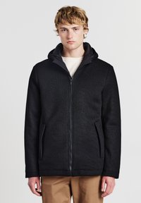 Giovane uomo con capelli biondi che indossa una giacca nera con zip e pantaloni beige, in piedi contro uno sfondo bianco semplice.