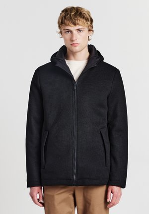 Giovane uomo con capelli biondi che indossa una giacca nera con zip e pantaloni beige, in piedi contro uno sfondo bianco semplice.