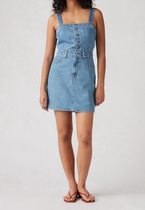 Kobieta w niebieskiej, bezrękawnikowej, zapinanej na guziki mini sukience z denimu, zestawionej z czerwonymi, płaskimi sandałami na cienkich paskach, na jednolitym tle.