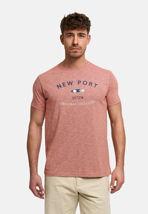 Mand iført en koral crewneck T-shirt med teksten "NEW PORT Original Indicode" og en lille grafik, kombineret med beige bukser.