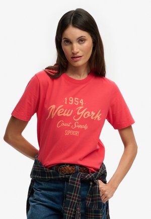 Mujer con camiseta roja "New York 1954 Coast Supply Superdry", jeans azules, cinturón trenzado marrón y una camisa de cuadros atada alrededor de la cintura.