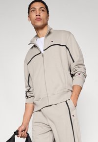 Μπεζ φούτερ με φερμουάρ και μαύρες γραμμές λεπτομέρειας, ριπ μανσέτες και λογότυπο στο στήθος. Συνδυασμένο με αντίστοιχο παντελόνι φόρμας. Ουδέτερη υφή υφάσματος.