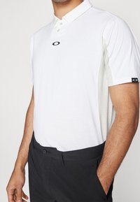 Polo shirt blanc à manches courtes avec col, présentant un logo noir sur la poitrine et des accents gris texturés sur les côtés.