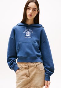 Blå kort hoodie laget av myk stoff, med en frontlomme og hvit logo-trykk. Kombinert med en beige denimskjørt.