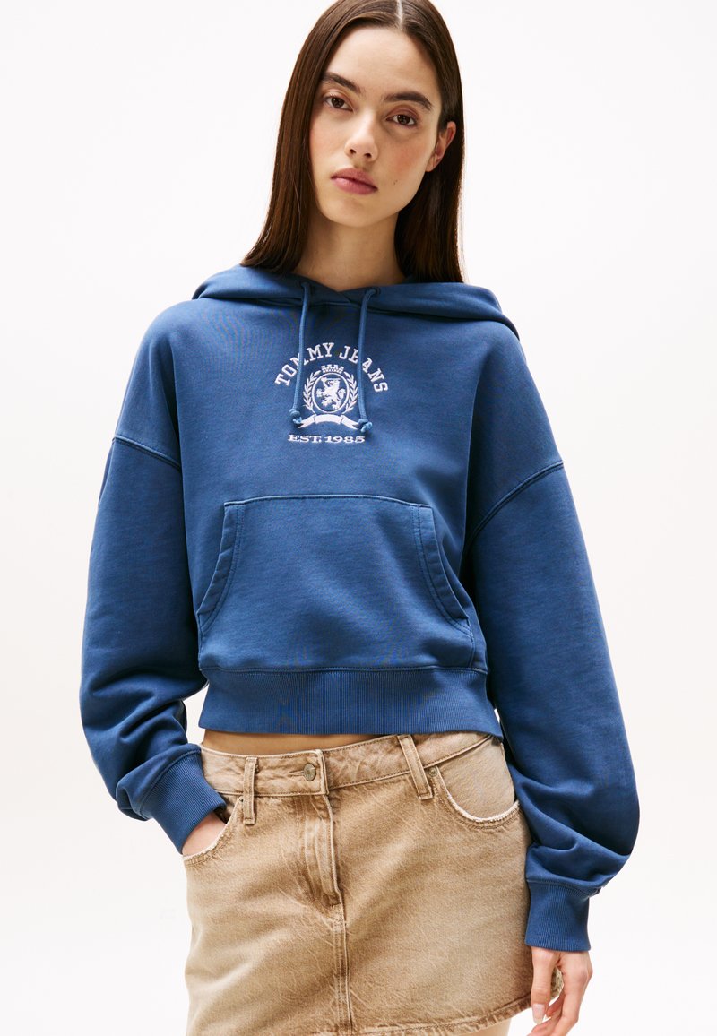 Blå kort hoodie laget av myk stoff, med en frontlomme og hvit logo-trykk. Kombinert med en beige denimskjørt.