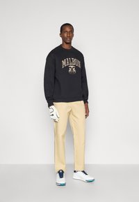 Melnā svītrā ar uzrakstu "MALBON" zeltītā krāsā, beigu bikses, un baltas golfa kurpes. Baltā cimdi ir uz kreisās rokas, stāvot pret vienkāršu fona.