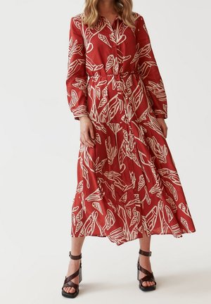 Vrouw draagt een rode jurk met lange mouwen met witte abstracte bladpatronen en zwarte sandalen met hoge hak en enkelbandjes.