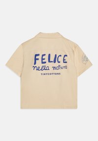 Camicia a maniche corte in cotone beige con testo blu "FELICE nella natura" e "TINY COTTONS" sul retro, con una toppa rotonda sulla manica.