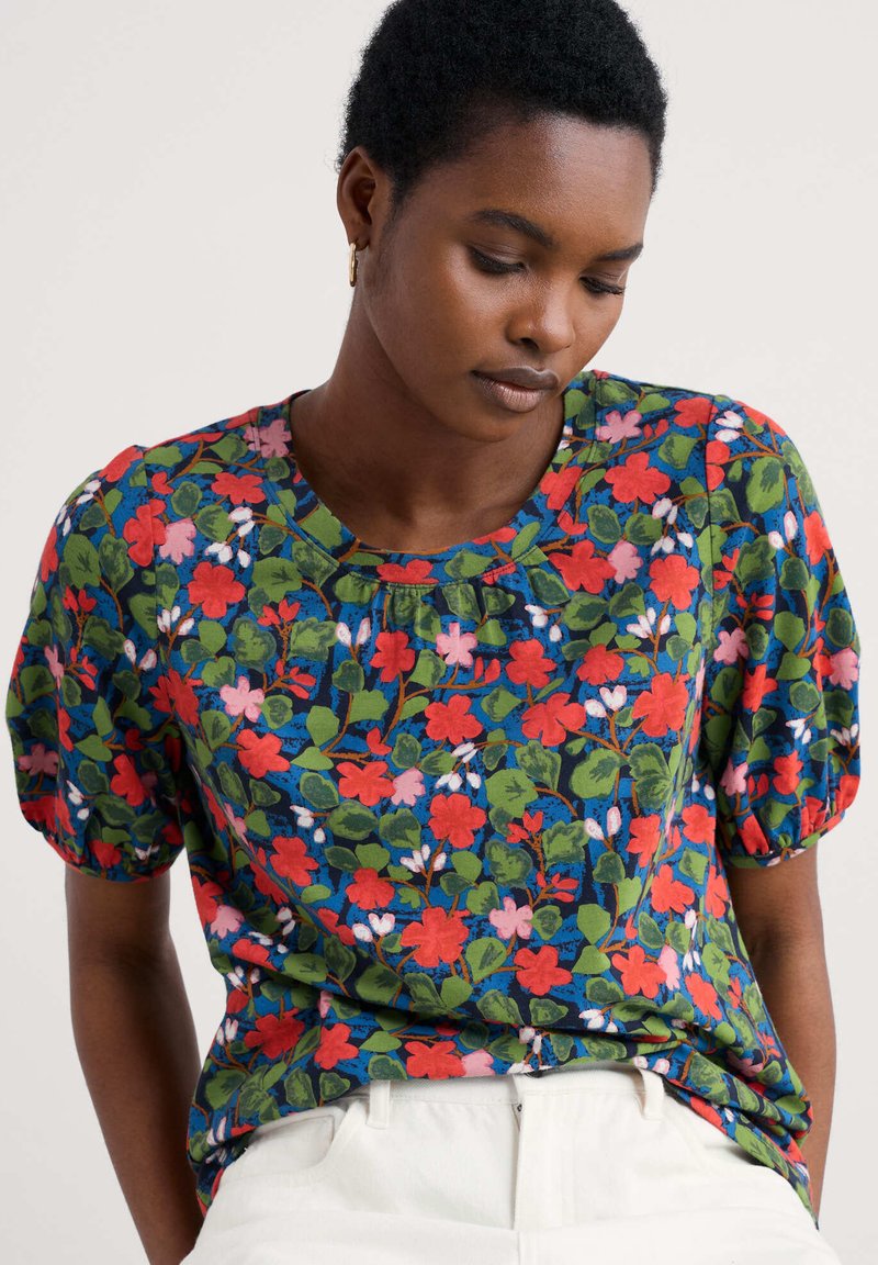 Blouse florale avec un fond bleu et des motifs rouges, roses et verts. Manches bouffantes et encolure ronde en tissu léger.