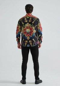Chemise à manches longues en tissu noir présentant un motif coloré et complexe avec des détails rouges, verts, bleus et dorés. Coupe ajustée et ourlet droit.