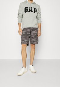 Felpa grigia con logo "GAP", pantaloni cargo camouflage e sneakers bianche. Focalizzazione su abbigliamento casual con una vestibilità rilassata e tasche pratiche.