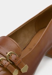 Καφέ δερμάτινο λόφερ με χρυσούς τόνους, featuring δύο κυκλικά αγκράφες και λείο φινίρισμα. Σχέδιο με επίπεδο τακούνι και στρογγυλή μύτη.