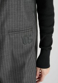 Zwart gestreepte stof met verticale lijnen; bevat een subtiele logo patch en textuur zwarte gebreide mouwen. Tailored design met zakken.