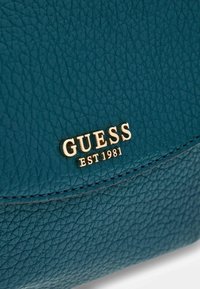 Torebka z tealowej skóry z fakturowaną powierzchnią, złotym logo "GUESS EST 1981" oraz strukturalnym kształtem z przeszyciami.