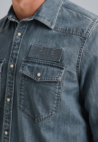 Niebieska koszula z denimu o zgaszonym kolorze, ze usztywnionym kołnierzykiem, podwójnymi kieszeniami na piersi, srebrnymi zatrzaskami oraz fakturowaną łatą z logo po lewej stronie.