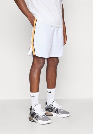 Baltas sporta šorti ar dzeltenām un melnām sānu svītrām, strukturēta auduma, apvienoti ar melnām un baltām sporta apaviem un baltām zeķēm.