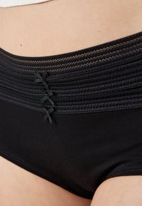 Slip in cotone nero con una vita testurizzata decorata da tre piccoli fiocchetti neri e dettagli a righe orizzontali sopra.