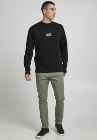 Zwarte sweatshirt met een witte grafische print op de borst, gecombineerd met lichtgroene broek en zwarte sneakers. De stof lijkt zacht en rekbaar te zijn.